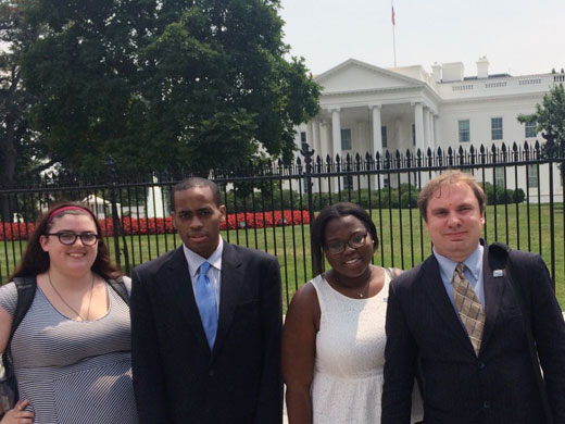 Four young professionals who are National Leadership Program participants - a white woman, a Black man, a Black woman and a white man - dressed professionally in suits standing in front of the fence in front of the White House