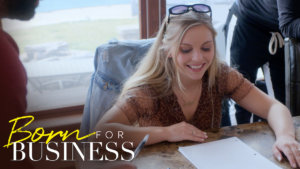 Lexi at a desk looking at a document smiling. Born for Business logo in bottom left.